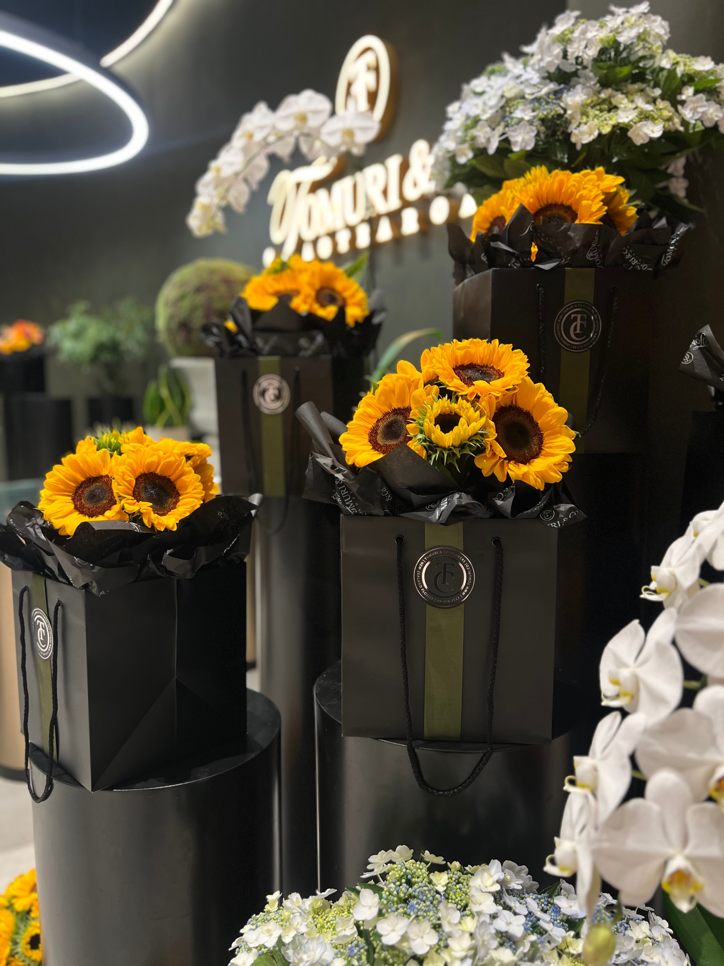 Floral arrangements with sunflowers in black gift bags against a dark background with 'Tomuri' branding.